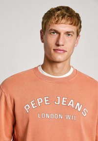 Sudadera de color melocotón con cuello redondo, cuello acanalado y texto en relieve blanco que dice "PEPE JEANS LONDON W11" en la parte frontal.