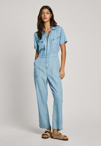 Light blue denim jumpsuit with short sleeves, front zipper, two chest pockets, tapered legs, and side pockets. Smooth texture, casual design.
