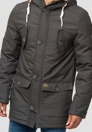Manteau d’hiver noir à capuche avec fermetures à boutons, deux poches poitrine à boutons et deux grandes poches avant à rabat, porté par une personne avec les mains visibles.