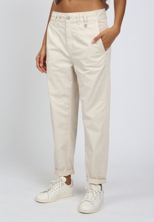 Beige Baumwollhosen mit hochgekrempelten Säumen, seitlichen Taschen und einem Knopfverschluss; getragen mit weißen Sneakers. Glatte Textur und entspannte Passform.