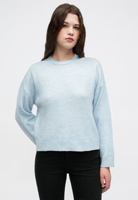 Lys blå strikket genser med rund hals, senkede skuldre og ribbestrikkede mansjetter og kant, kombinert med mørke jeans. Mykt og teksturert stoff.