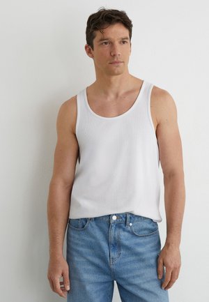 Homme portant un débardeur blanc sans manches et un jean bleu, debout contre un mur blanc, regardant légèrement vers la droite.