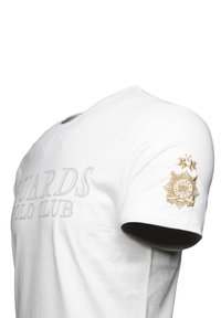 Camiseta de algodón blanca con un estampado texturizado "GUARDS POLO CLUB" y un emblema bordado dorado en la manga izquierda. Mangas cortas y cuello redondo.