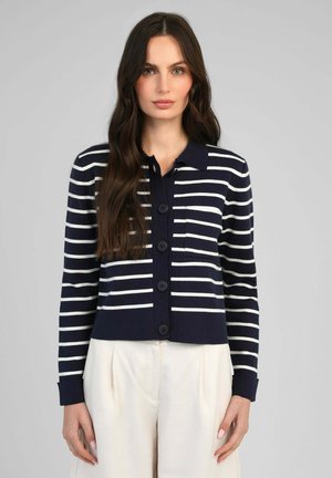 Femme aux longs cheveux foncés portant un cardigan boutonné rayé bleu marine et blanc ainsi qu’un pantalon blanc, debout devant un fond gris uni.