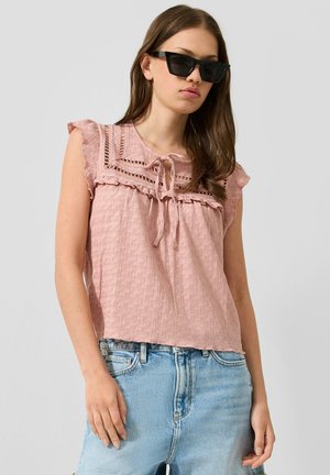 Junge Frau mit schwarzen Sonnenbrillen, einer rosa strukturierten ärmellosen Bluse mit Spitzendetails und hellblauen Jeans vor einem einfarbigen Hintergrund.