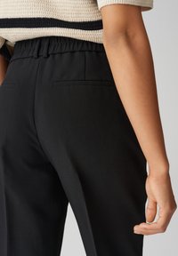 Pantalons noirs ajustés avec une texture lisse, ceinture élastique et deux poches arrière, présentés de côté avec une vue partielle d'un haut en tricot.