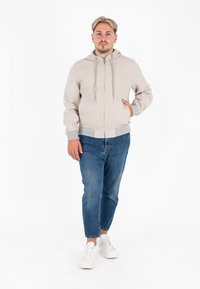 Felpa zip-up beige chiaro con cappuccio regolabile, due tasche frontali, polsini a coste, abbinata a jeans blu e sneakers bianche. Tessuto liscio.