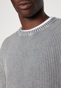 Harmaa neulottu villapaita ribbineuloksisella kaula-aukolla, jossa on tekstuurillinen kangas ja päällekkäinen valkoinen paita alla.