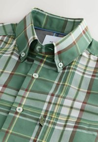 Chemise à carreaux verts, bruns, blancs et jaunes avec col à boutons et étiquette de marque visible à l'intérieur du col et sur la couture latérale.