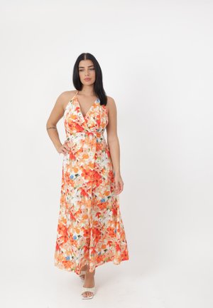 Femme aux longs cheveux noirs portant une robe maxi sans manches orange et blanche à fleurs et des talons blancs ouverts, debout devant un fond blanc.