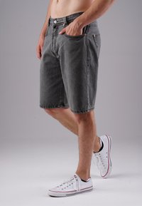 Man wearing gray denim shorts and white sneakers stands with one hand in pocket against plain gray background.
