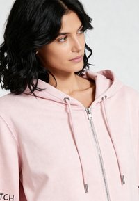 Roza dukserica na patentni zatvarač s kapuljačom i vrpcama za vezanje, izrađena od mekog tkanog materijala, sadrži srebrne metalne detalje i suptilnu teksturu.