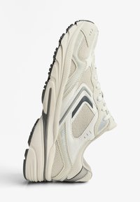 Béžová športová obuv s sieťovinou a syntetickým materiálom, zaoblený tvar, textúrovaný dizajn, biele šnúrky a tmavá gumová podrážka s drážkami.