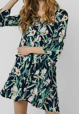 Femme portant une robe fleurie foncée avec des fleurs blanches et des feuilles vertes, avec des manches longues et une jupe à volants.