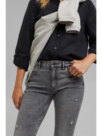 Camisa preta de botão, suéter cinza pousado sobre os ombros e jeans cinza de cintura alta com desgaste e corte justo.