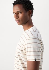 Striped short-sleeve t-shirt in cream with brown zigzag lines, featuring a crew neck and a small black label on the left sleeve.
