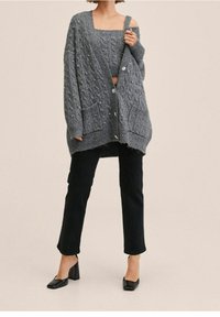 Grau gestrickte Strickjacke mit strukturiertem Zopfmuster, vorderen Taschen und Knöpfen, getragen über einem passenden, kurz geschnittenen Oberteil und kombiniert mit schwarzen Jeans und Absätzen.