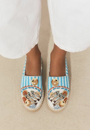 Feet wearing blue and white striped slip-on shoes decorated with colorful beaded seafood and citrus fruit designs.