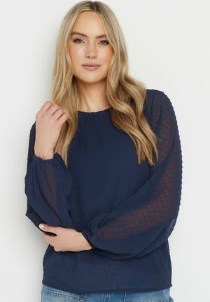 Navyblauwe blouse met gestructureerde stippen, ronde halslijn en doorschijnende pofmouwen. Gecombineerd met denim jeans.