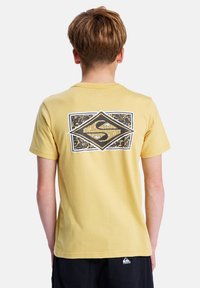 Žluté bavlněné tričko s krátkým rukávem, které na zadní straně zdobí texturovaný grafický motiv ve tvaru diamantu v černé a bílé barvě se designovými detaily.