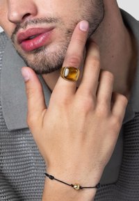 Guldring med en stor, rektangulær ravsten, båret på hånden med fingrene berørende hagen. Sort snorearmbånd med en guldaccentperle.