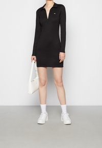 Black long-sleeve dress with a collared neckline and a front placket. Accompanied by a white handbag and white platform sneakers.
