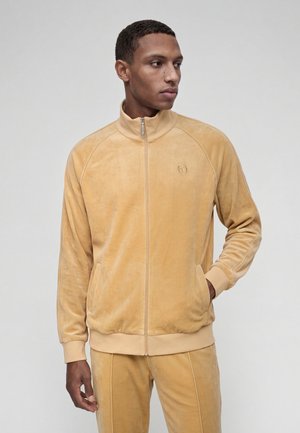 Homme portant une veste zippée en velours marron clair et un pantalon assorti, debout, une main dans la poche, regardant sur le côté.