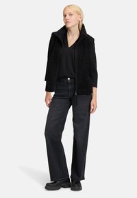 Czarna bezrękawnikowa kamizelka z fakturowanej tkaniny, w połączeniu z czarną bluzką i ciemnymi dżinsami. Model stoi w czarnych butach na grubej podeszwie.