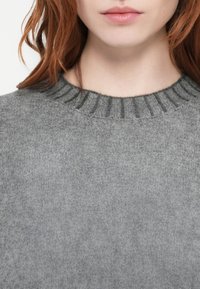 Grå strikkegenser med ribbede kanter. Teksturen er myk med et subtilt, jevnt mønster. Stoffet ser varmt og koselig ut.