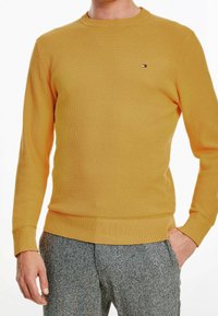 Maglione giallo a maglia con collo rotondo, polsini e orlo a coste. Presenta un piccolo logo sul petto. Indossato con pantaloni grigi con trama.