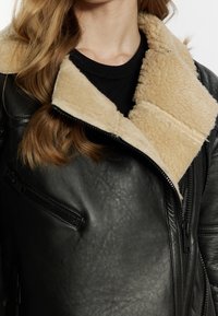 Giubbotto di pelle nera con colletto in ecopelliccia beige, dotato di tasche con zip e una finitura testurizzata.