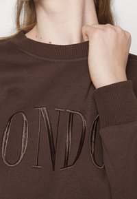 Brun sweatshirt med ribbad halsringning och muddar, med präglad text som stavar "LONDON" i matchande satinfinish. Mjukt tyg.