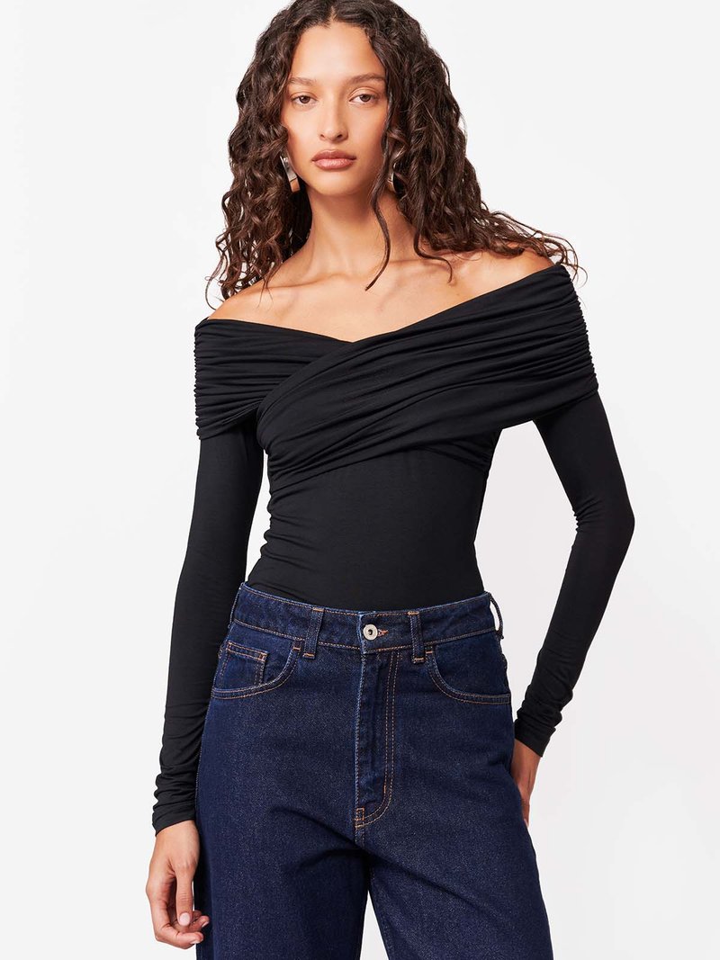 Volüümikate lokkis juustega naine, kes kannab musta ühe-õlaga pika varrukaga pusa ja tumedates sinistes kõrge vöökohaga teksapükse valge tausta ees.