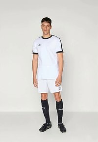 Vīrietis stāv tērpies baltā sporta krekla un šortos, melnās līdz ceļiem garās zeķēs un melnās kurpes, pret vienkāršu gaišu fona.