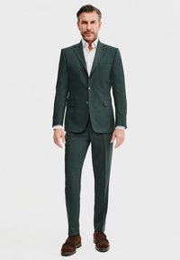 Ciemnozielony garnitur z dopasowanym krojem, jednorzędowa marynarka i dopasowane spodnie. Biała koszula w kropki. Brązowe buty.