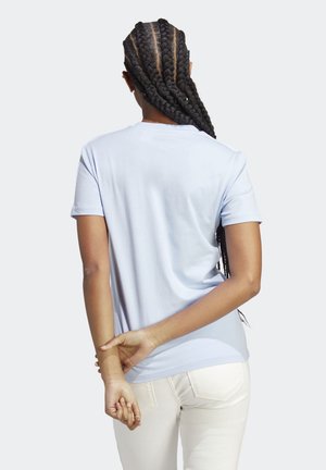 Vrouw met lang gevlochten haar, gekleed in een lichtblauw T-shirt en witte broek, staand met haar handen achter haar rug gevouwen.