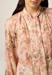Blusa rosa chiaro con motivo floreale in tonalità di verde e rosa. Design plissettato con collo alla americana e texture di tessuto morbido e leggero.