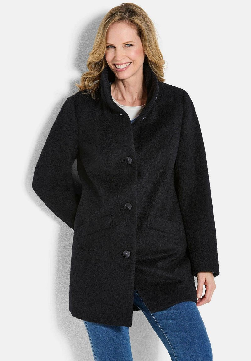 Cappotto nero con texture e colletto alto, caratterizzato da cinque bottoni, tasche laterali e un taglio dritto. Indossato sopra un top bianco.