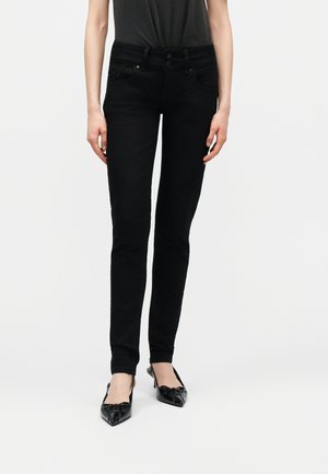 Mujer con vaqueros ajustados negros y zapatos de tacón con punta en pico y tira trasera, de pie frente a un fondo claro y liso.