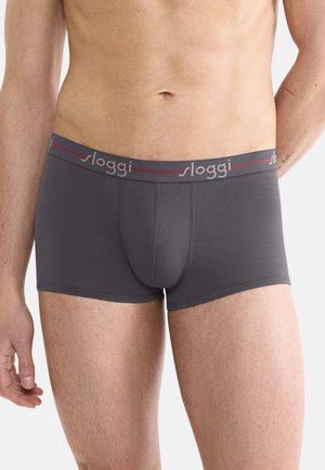 Mand iført grå Sloggi boxershorts med en logo på taljebåndet, stående mod en ensartet hvid baggrund.