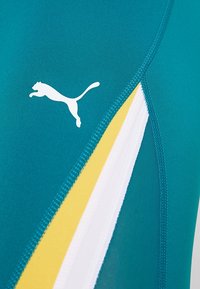 Niebieska bluzka sportowa z białym logo Puma. Boczne panele z żółtymi i białymi paskami, gładka tekstura oraz dopasowany krój.