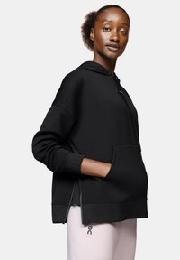 Sweat-shirt à capuche noir avec une poche kangourou, des poignets côtelés et des zips sur les côtés. Texture douce avec une coupe décontractée, associé à un pantalon léger.