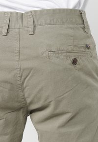 Close-up van beige textuur broek met een knoopzak aan de achterkant en riemlussen, gedragen met een ingestopt wit shirt.