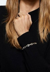 Silberne Kettenarmband mit runden Gliedern, am Handgelenk getragen, kontrastiert mit der Textur und Farbe eines schwarzen Pullovers. Lange, glatte braune Haare sind sichtbar.