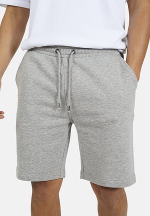 Uomo che indossa pantaloncini grigi chiaro con coulisse e mani nelle tasche laterali, con una maglietta bianca.