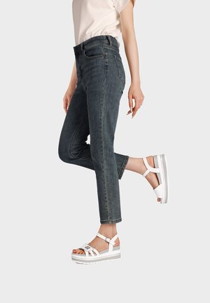 Persona con jeans slim scuri e sandali platform bianchi, una gamba piegata all’indietro su uno sfondo chiaro e uniforme.