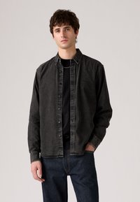 Black denim shirt with a button-up design, pointed collar, and contrast stitching, worn over a black t-shirt, paired with dark jeans.