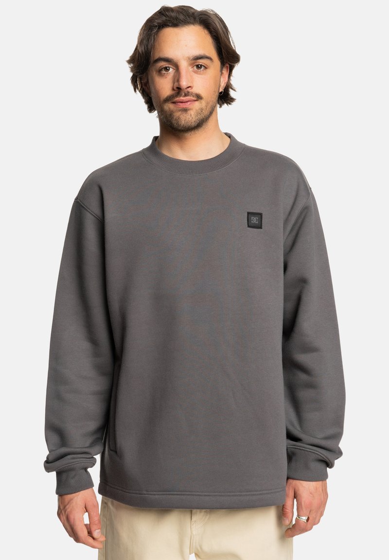 Sudadera gris con cuello redondo, puños acanalados y un parche rectangular negro con logo en el lado izquierdo del pecho. Textura suave, corte relajado.