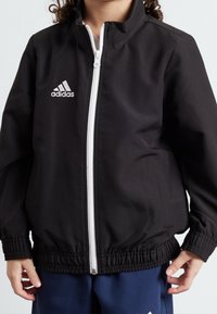 Chaqueta negra con cremallera hecha de un tejido suave; presenta una cremallera blanca y el logo de Adidas en el lado izquierdo del pecho; tiene un cinturón y puños elásticos.