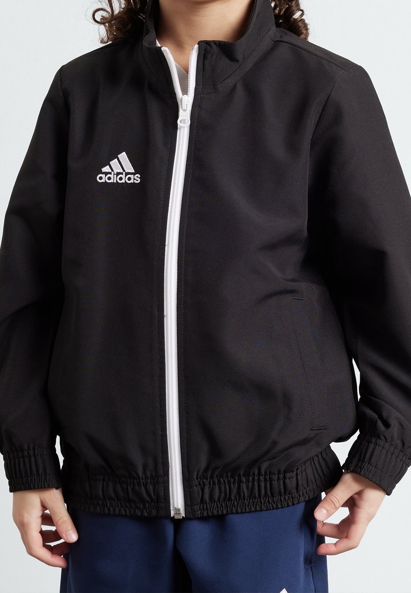 Chaqueta negra con cremallera hecha de un tejido suave; presenta una cremallera blanca y el logo de Adidas en el lado izquierdo del pecho; tiene un cinturón y puños elásticos.